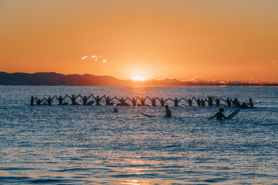 2022年の元旦に茅ヶ崎ヘッドランドビーチの海上で手をつないで新年を祝うサーファーたちと海と空を赤く染める初日の出